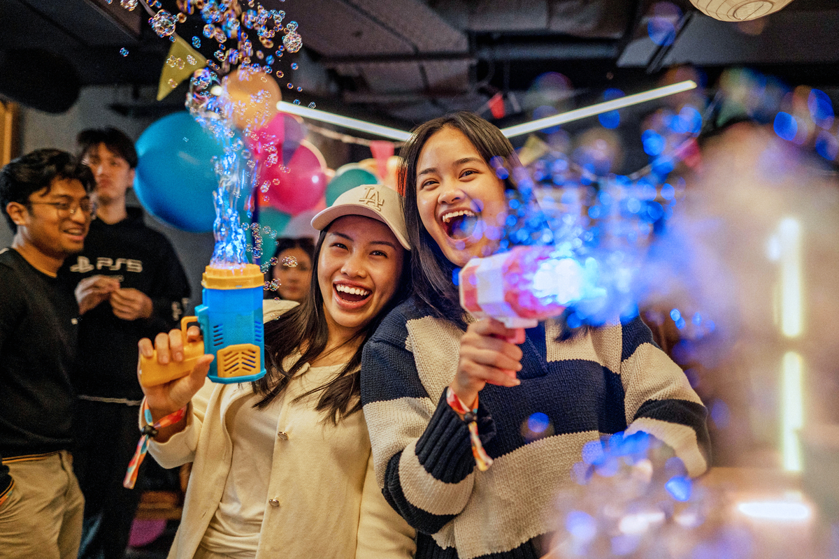 Two smiling women wearing casual clothes are playfully shooting bubble guns, surrounded by colorful bubbles and balloons. The vibrant atmosphere suggests a festive or party setting, with blurred figures and lights in the background.